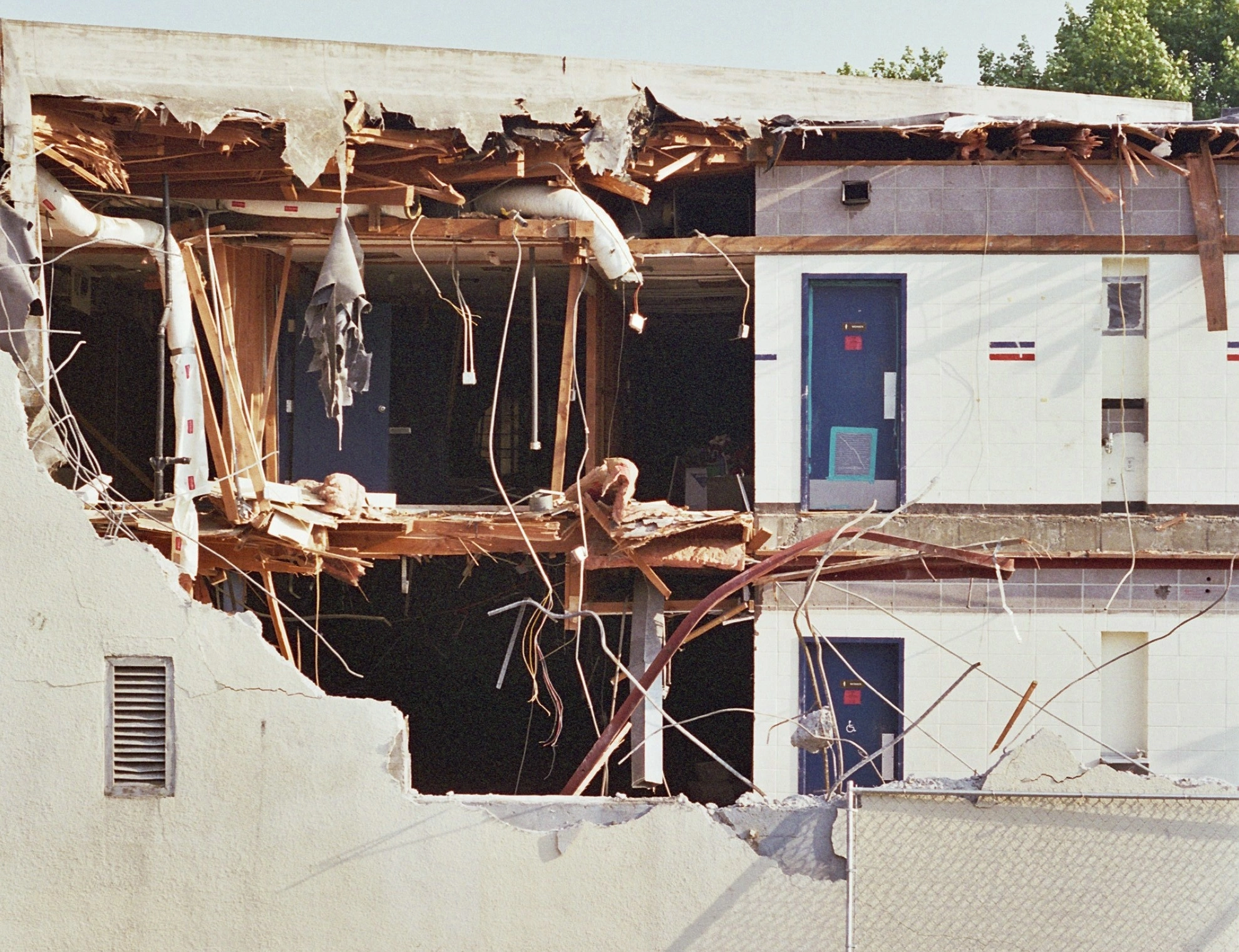 Partially demolished multi-storey building in Singapore showing exposed wooden framing, hanging wires, broken concrete walls, remaining blue doors, and construction debris behind a temporary fence.