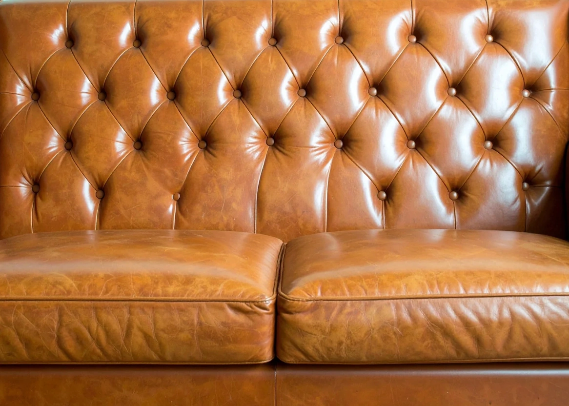 Close-up of a full-grain leather sofa in rich caramel brown, showing deep diamond button tufting on the backrest and soft, naturally creased seat cushions that highlight the leather’s texture and durability.